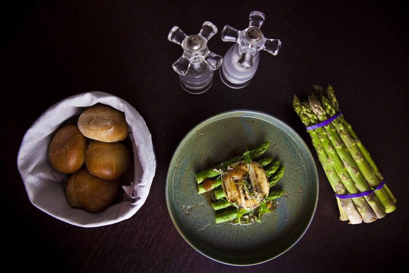 Asparagus dish in a green bowl beside a bunch of asparagus, bread rolls and oil cruets, from above