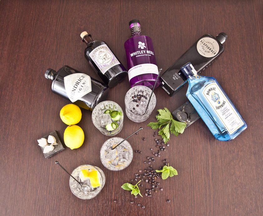 Gin bottles, lemons and glasses of ice laid out on a dark table
