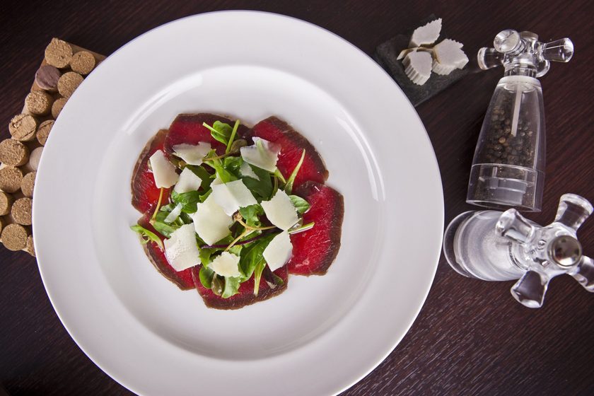 Beef carpaccio with rocket and parmesan shavings on a white plate, corks and a pepper mill beside it