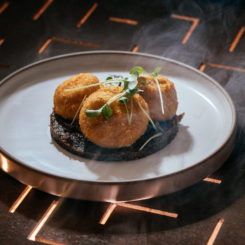 Three crumbed baked sheep's cheese croquettes on a dark fig chutney, steaming, on a copper lit table